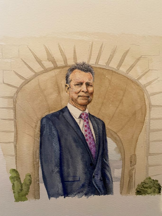 A man in a suit stands under an archway with a smiling expression.